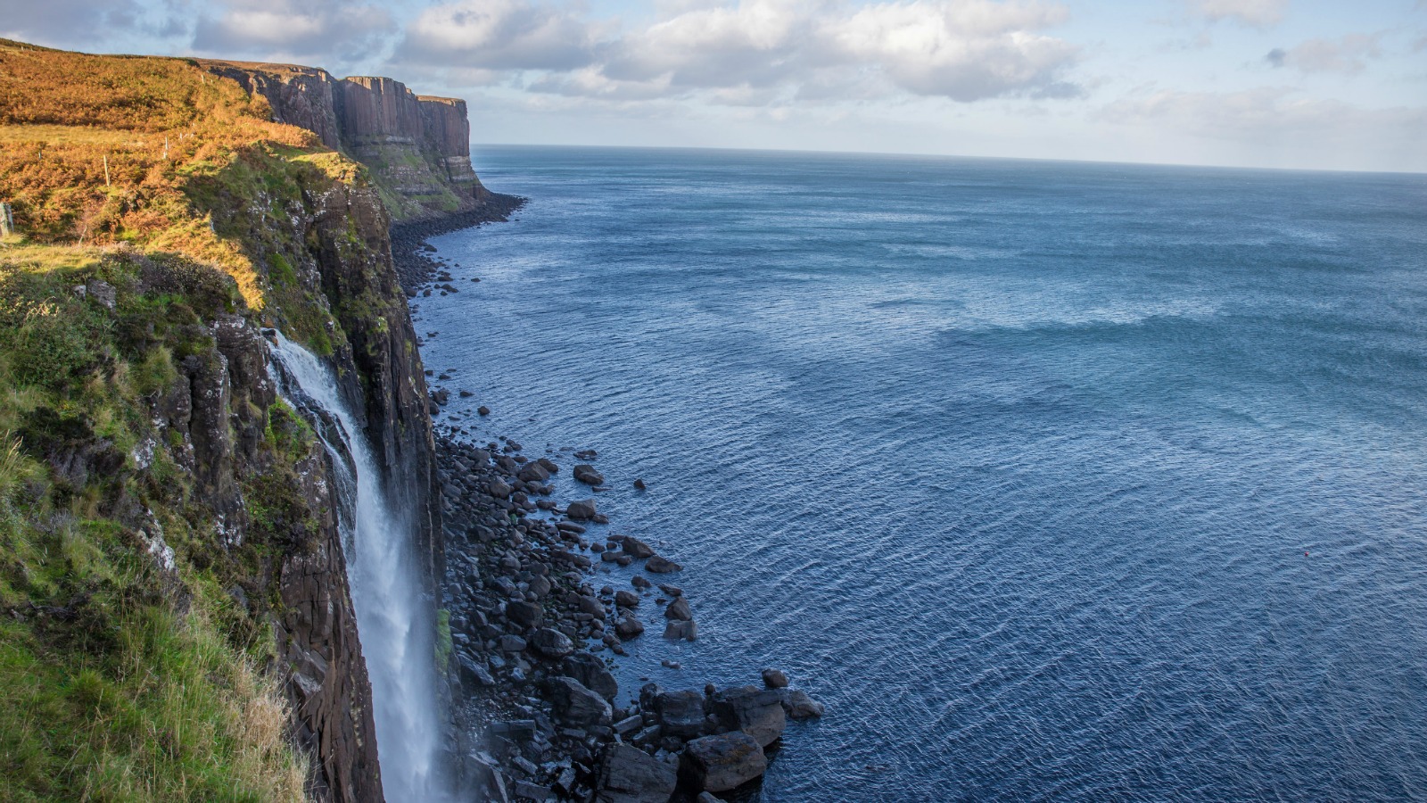 Kilt Rock Waterfall cascading from the cliffside on a sunny day, with bright skies and coastal views